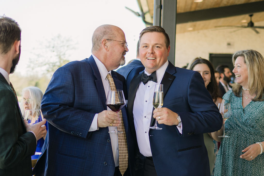 A groom smiles while raising a toast, surrounded by loved ones and champagne flutes in this joyful wedding reception moment.