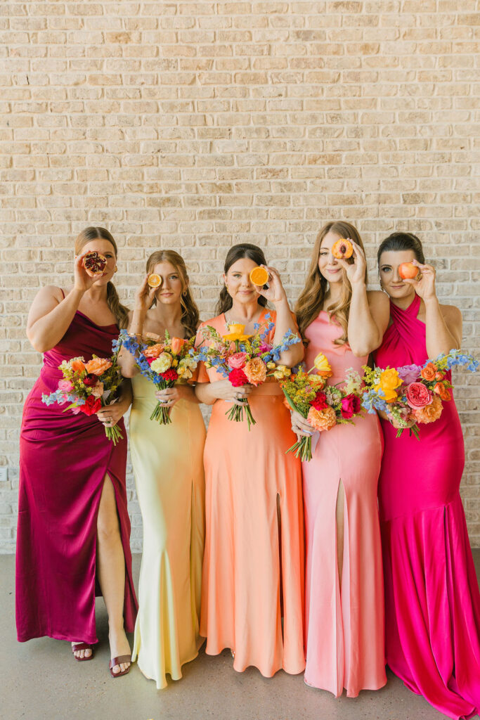 A group of bridesmaids posed while holding fruit against the neutral brick backdrop of this modern Texas wedding venue.