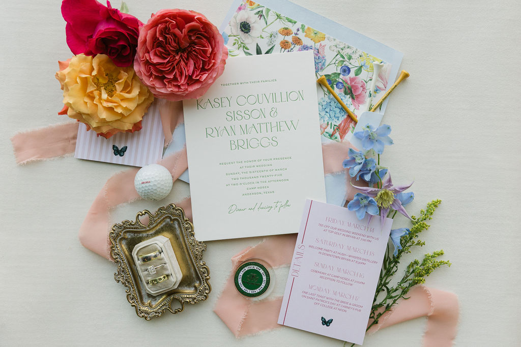A flat lay of the couple’s invitation suite, featuring floral patterns, a large pink bloom, and golf-themed details that reflect their unique personality and wedding theme at Camp Hosea.