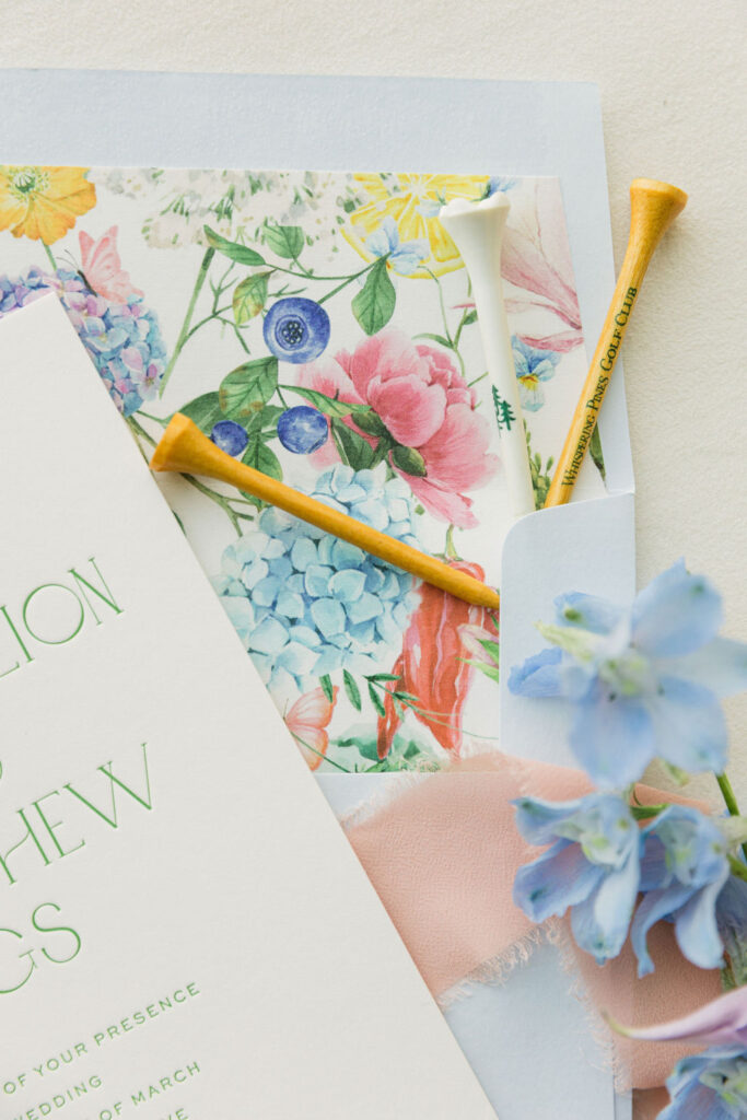 A close-up flat lay of the RSVP card and floral envelope liner with yellow golf tees and baby blue delphinium, tying in colorful details from this Houston wedding day.
