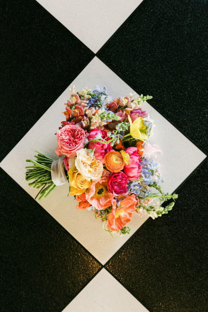 A vibrant bouquet with ranunculus, poppies, roses, and delphinium rests on the black-and-white checkered floor of Camp Hosea’s reception space.