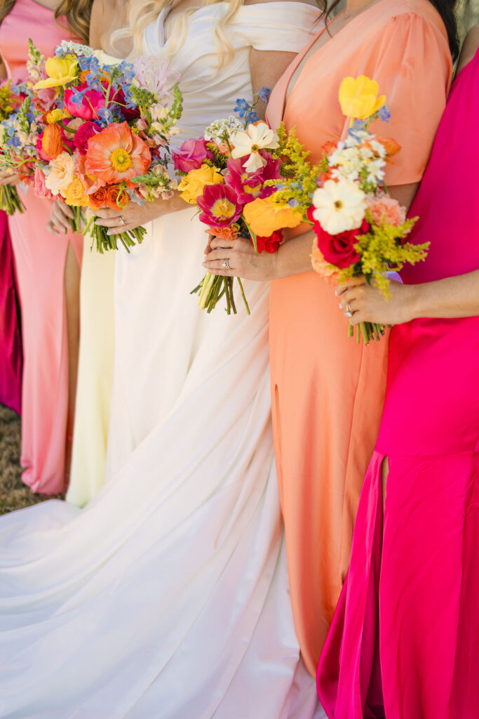 Bridesmaids in vibrant coral, fuchsia, and orange gowns hold colorful bouquets beside the bride, styled to perfection for a Camp Hosea wedding.