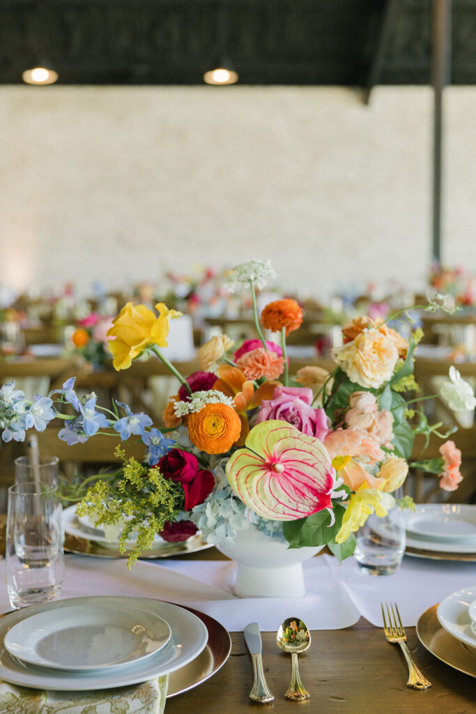 A colorful floral centerpiece with ranunculus, anemones, and roses sits on a wooden reception table at Camp Hosea, ideal for couples wanting colorful wedding inspiration.