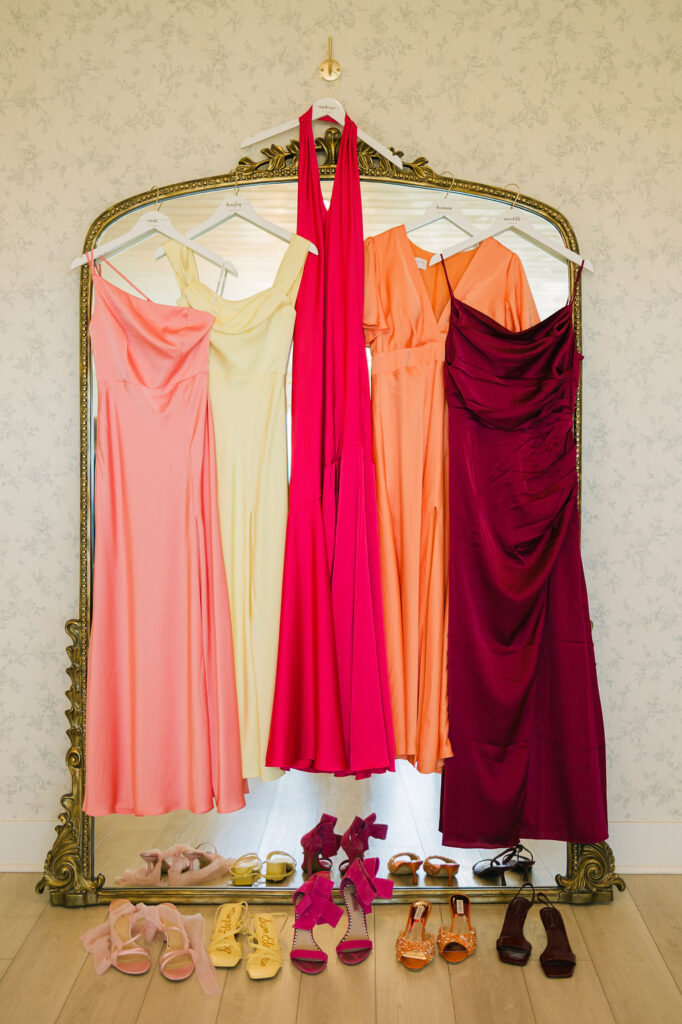 Bridesmaid dresses in bold tones of red, orange, and yellow hang from a gold mirror, with coordinating shoes displayed underneath—a vibrant color story for this Camp Hosea wedding.