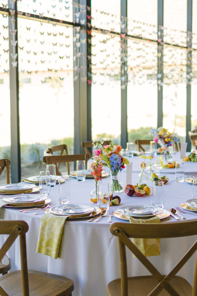 Reception tables set with gold napkins, colorful florals, and modern chairs line the glass walls of Camp Hosea’s light-filled dining space.