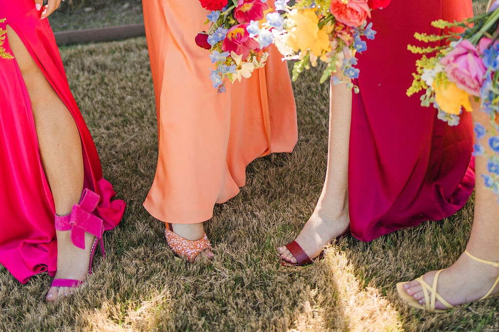 A detail of colorful wedding shoes in orange and burgundy peeking out from bridesmaid dresses on the lawn outside Camp Hosea.