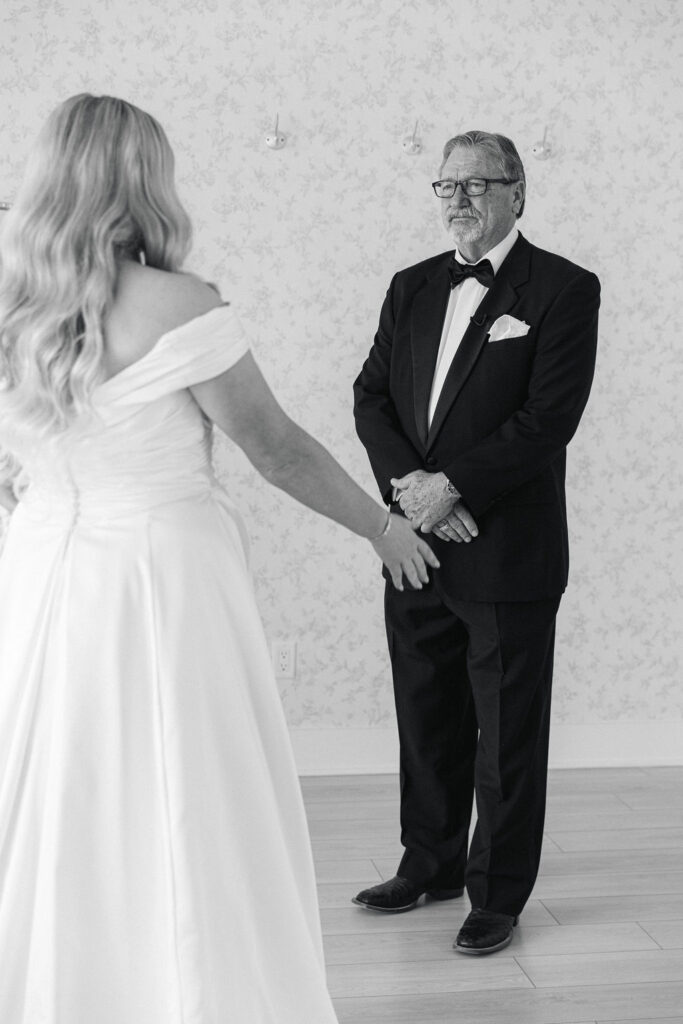 The bride sees her father during their first look, captured in soft lighting with a wallpapered bridal suite as the backdrop.