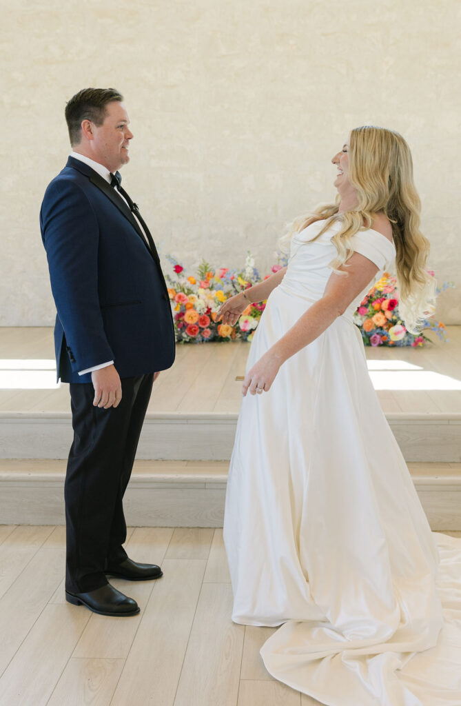 The couple laughs during their first look in the chapel at Camp Hosea, framed by bright natural light and pastel florals.
