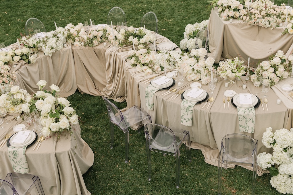 Elegant outdoor wedding tables arranged in a S-shape with taupe linens, romantic white florals, and transparent ghost chairs on the lawn