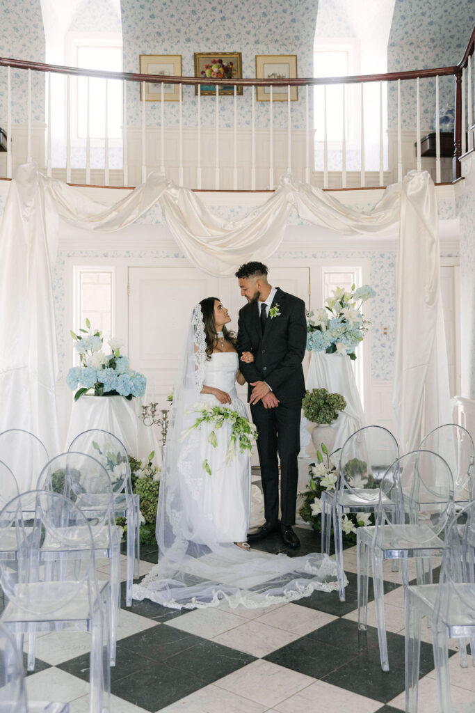 Bride and groom stand arm-in-arm in an elegant indoor ceremony setup at Greystone Castle, surrounded by white lilies, blue hydrangeas, and ivory draping.