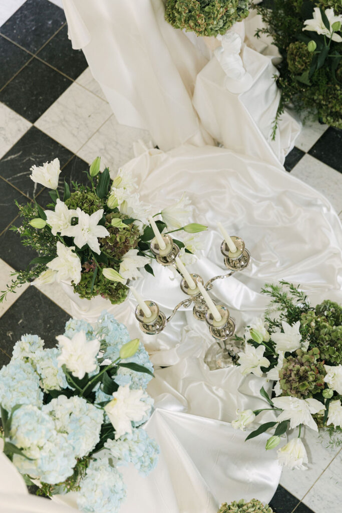 Overhead view of an elegant indoor ceremony setup at Greystone Castle with white lilies, blue hydrangeas, lush greenery, satin draping, and a silver candelabra on black and white checkered tile.