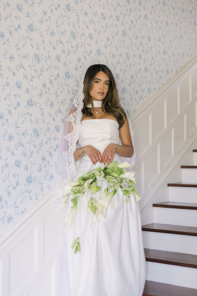 Bride poses on a staircase lined with white paneling and vintage floral wallpaper at Greystone Castle