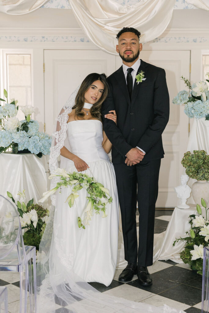 Bride and groom stand arm-in-arm in an elegant indoor ceremony setup at Greystone Castle, surrounded by white lilies, blue hydrangeas, and ivory draping.
