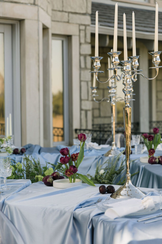 greystone castle outdoor wedding reception table set up with blue draped linen, green florals, Brussels sprouts garland, dark plums, and tall taper candles in silver holders.