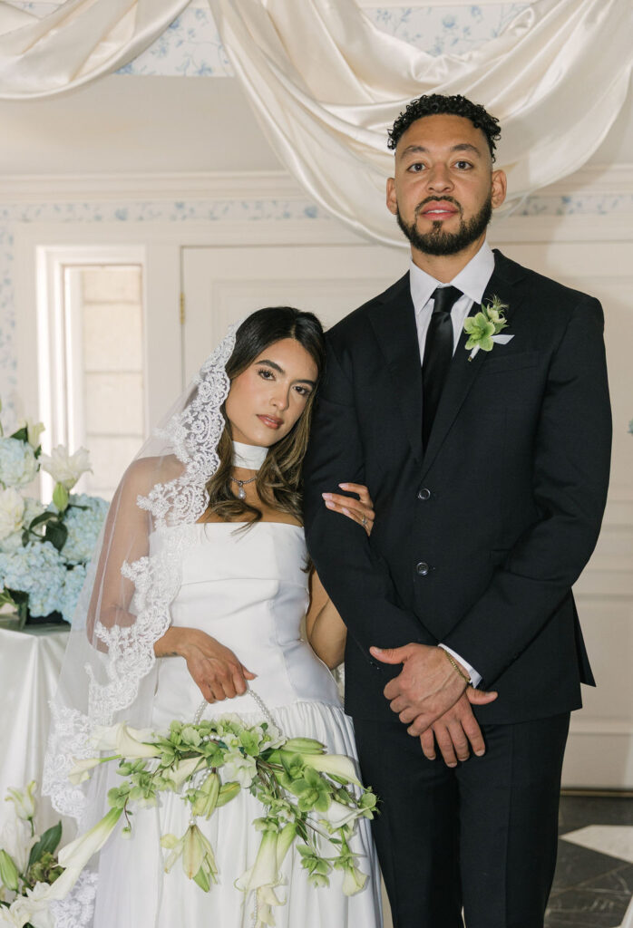 Bride and groom stand arm-in-arm in an elegant indoor ceremony setup at Greystone Castle, surrounded by white lilies, blue hydrangeas, and ivory draping.