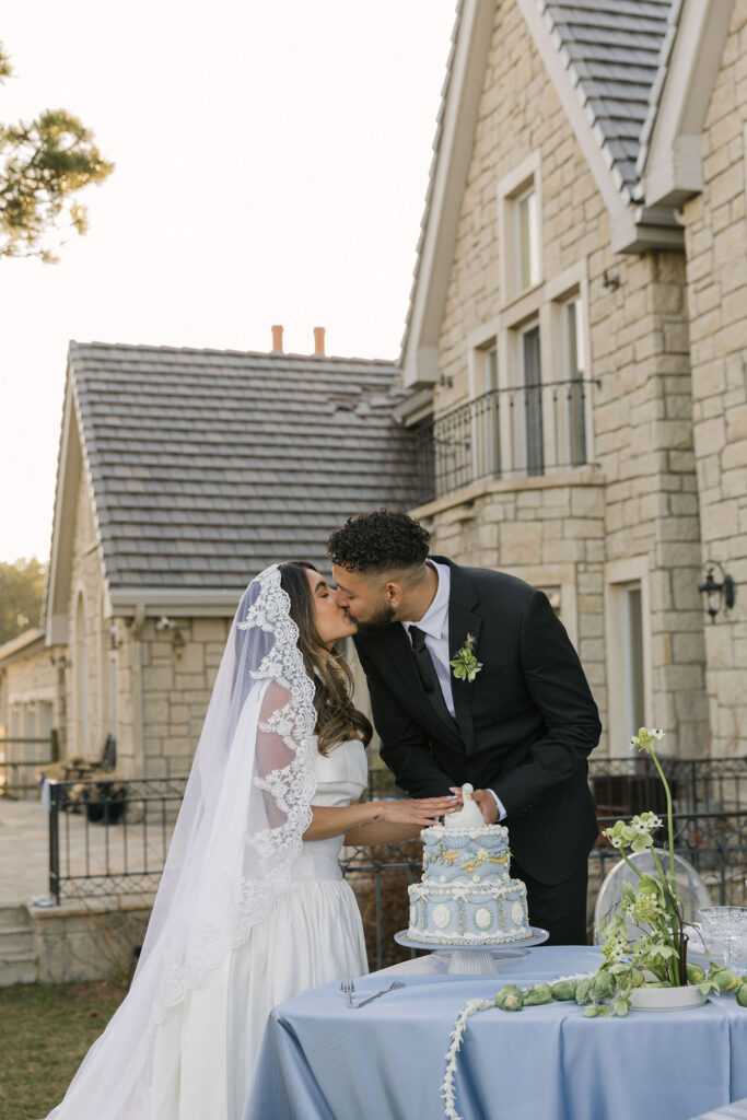 bride and groom kissing while cutting a vintage-inspired blue wedding cake topped with a porcelain swan, set against the stone backdrop of Greystone Castle.