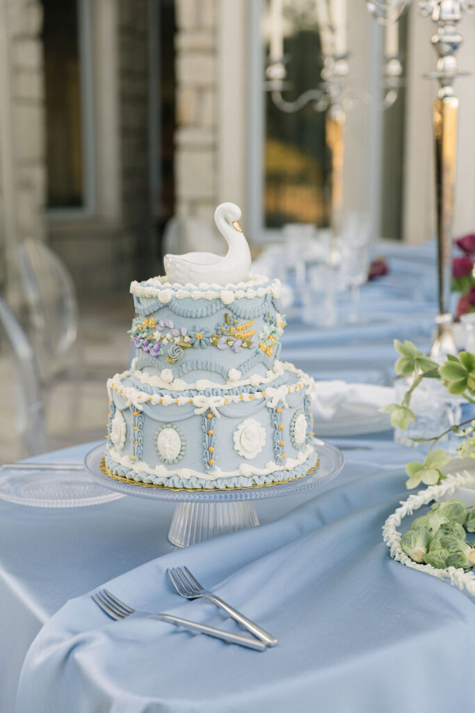 Whimsical two-tiered wedding cake with ornate blue and white piping, cameo details, and a swan topper