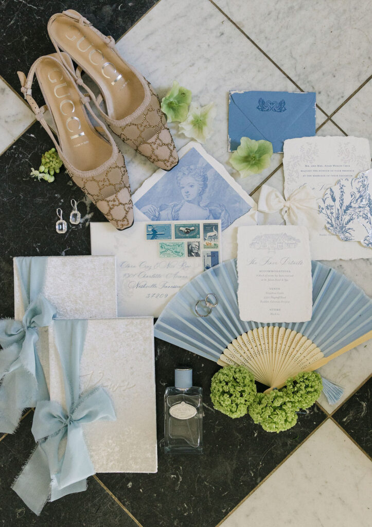 Romantic flat lay featuring vintage stamps, blue-toned fine art invitations, wedding rings, delicate green flowers, a paper fan, and sparkly Gucci bridal shoes.