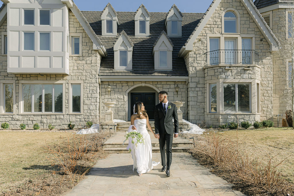 Bride and groom walk hand-in-hand outside Greystone Castle, the stone exterior and dramatic architecture framing their joyful exit in golden afternoon light.