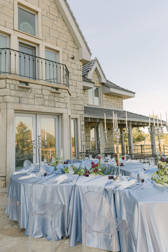 greystone castle outdoor wedding reception table set up with blue draped linen, green florals, Brussels sprouts garland, dark plums, and tall taper candles in silver holders.