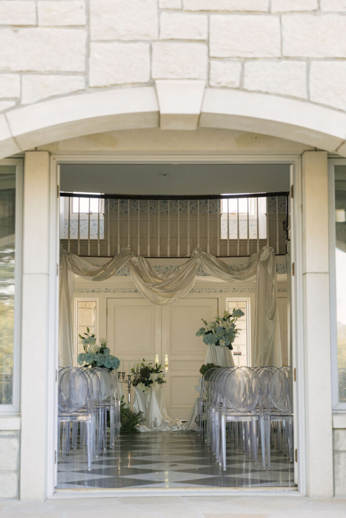 Elegant view through arched stone doors into the indoor wedding ceremony setup at Greystone Castle, featuring soft ivory drapery, ghost chairs, and romantic blue-and-white florals.