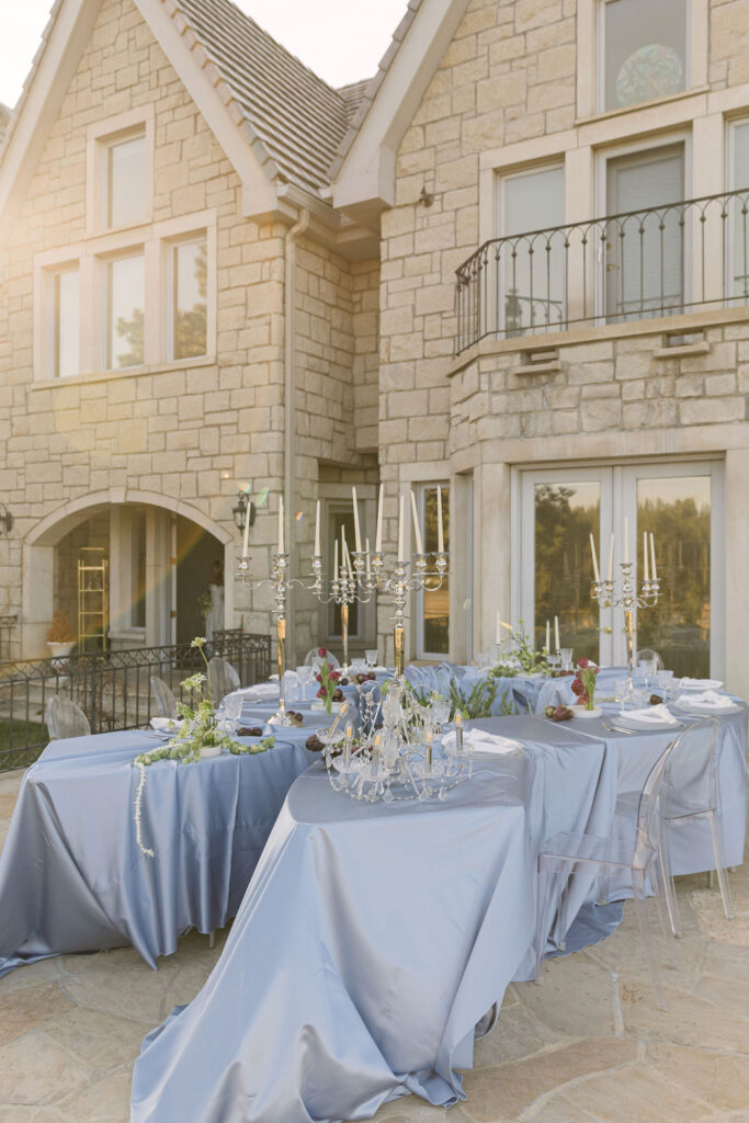 Greystone Castle wedding reception set up outdoors, with blue-draped wedding reception table decorated with green florals, Brussels sprouts garland, dark plums, and tall taper candles in silver holders.