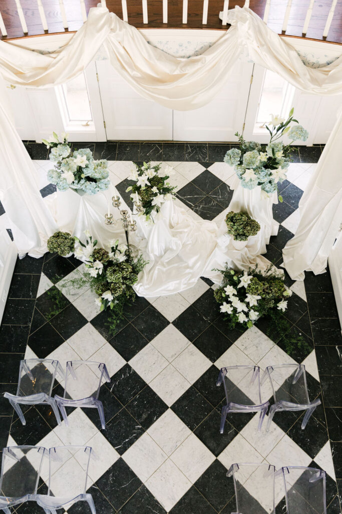 Overhead view of an elegant indoor ceremony setup at Greystone Castle with white lilies, blue hydrangeas, lush greenery, satin draping, and a silver candelabra on black and white checkered tile.