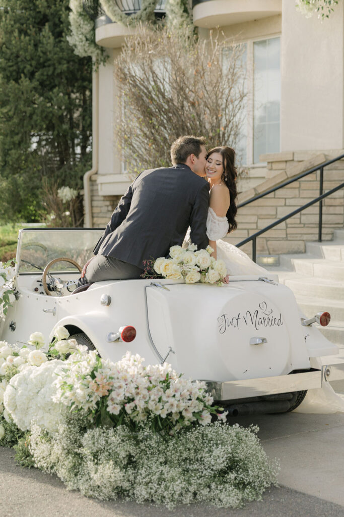 Bride and groom share a sweet moment in a vintage white convertible decorated with flowers and a “Just Married” sign in front of Chateaux at Fox Meadows, a wedding venue in Broomfield, CO