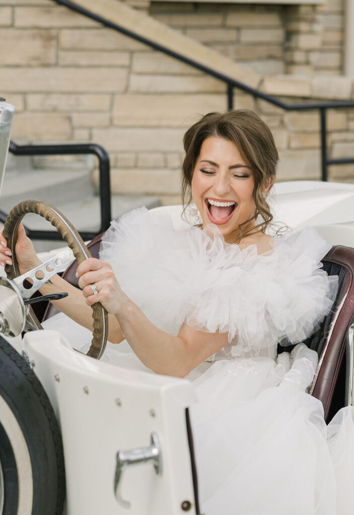 Bride sits joyfully in a vintage white convertible surrounded by lush floral arrangements outside Chateaux at Fox Meadows