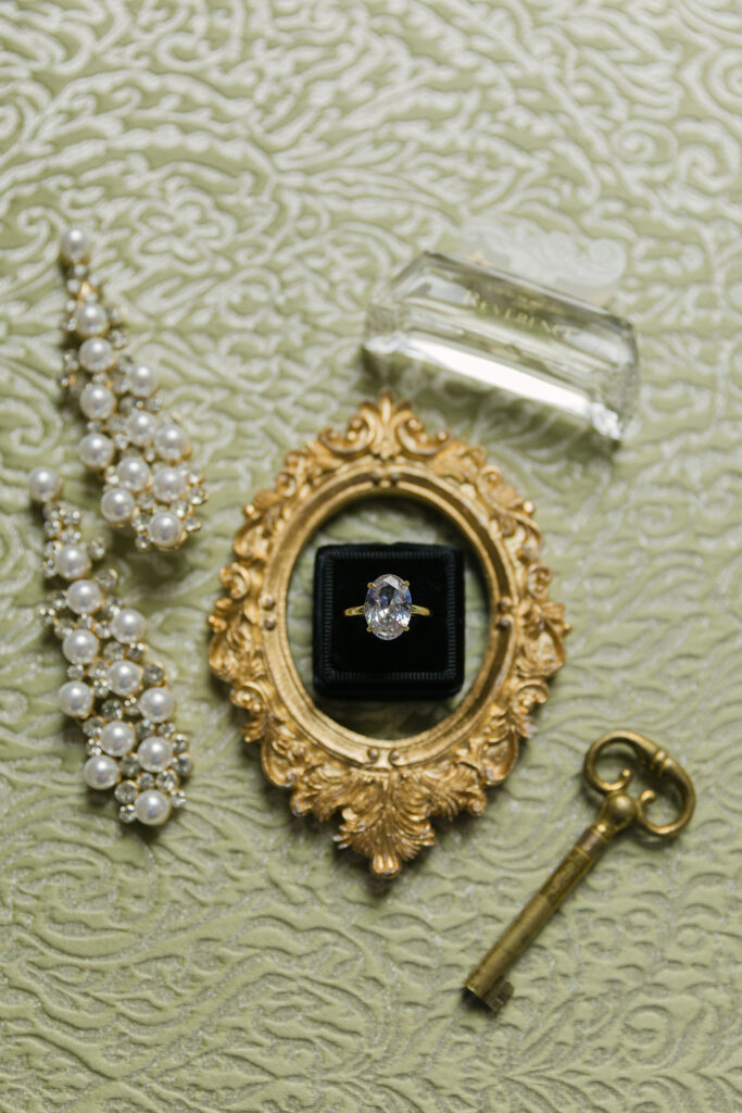 Romantic bridal flat lay with an oval engagement ring in a black velvet box, vintage gold frame, pearl earrings, perfume, and an antique key on a green textured fabric.