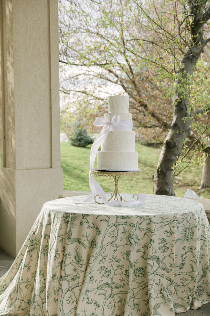A four-tier white wedding cake tied with a satin ribbon displayed on a floral-printed tablecloth with trees in the background