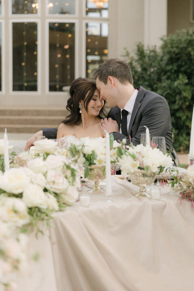 The groom gently kisses the bride’s forehead as they sit at a candlelit sweetheart table surrounded by ivory blooms