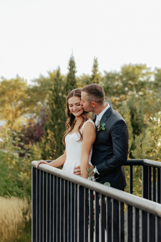 Groom kissing bride on the cheek on the balcony overlooking the lush garden ceremony site at Brookside Gardens Weddings and Events