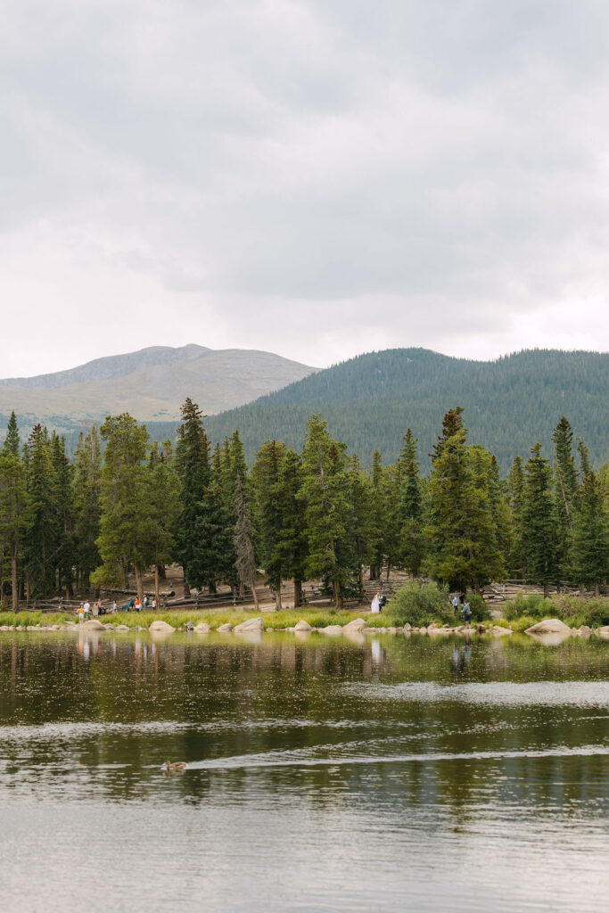 Bride and groom off in the distance exchanging private vows at Echo Lake among the mountains and sitting on a bench by the water 