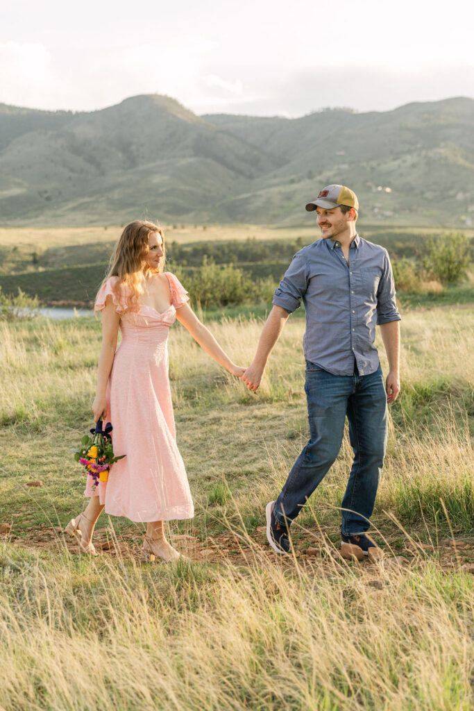 Couple holding hands and walking through tall grass during their sunset engagement session.