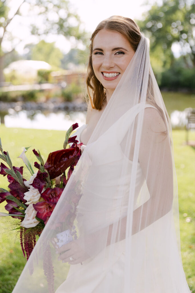 Bride smiling softly under her veil holding a deep-red bouquet during an elegant garden wedding at Hudson Gardens.