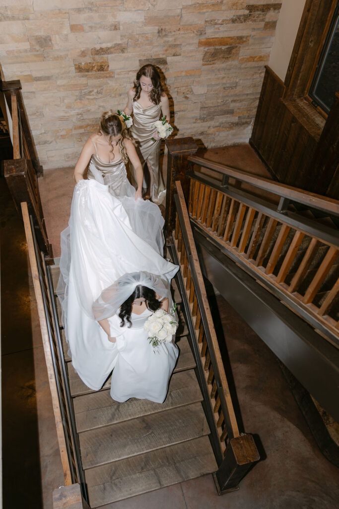 Candid wedding photo of bridesmaids helping the bride down the stairs inside Spruce Mountain Ranch wedding venue 