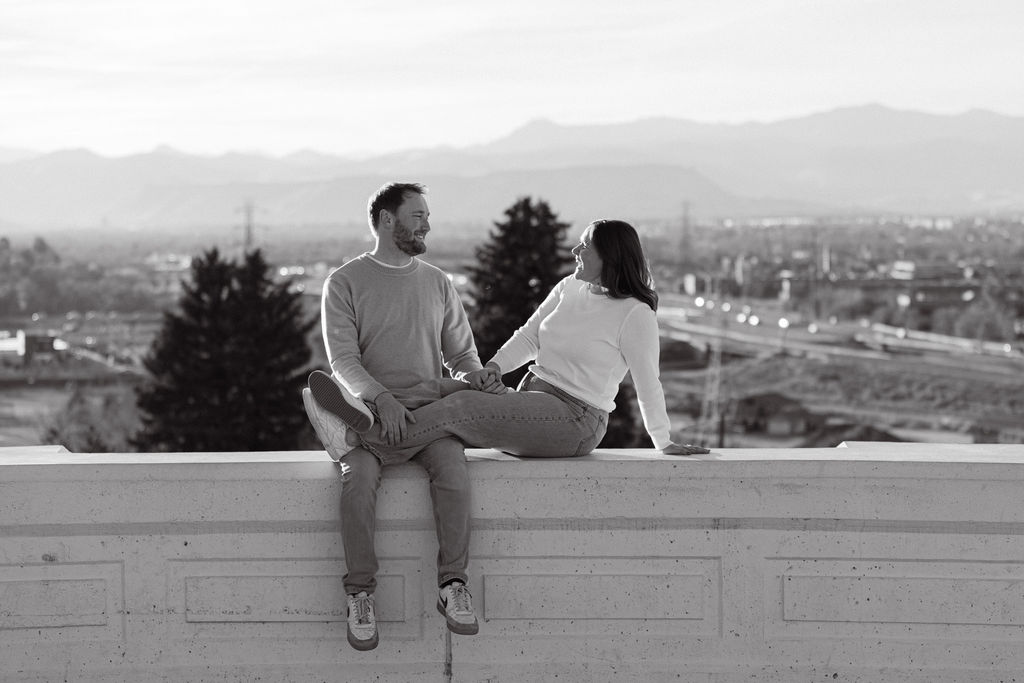 Black and white photo of couple sitting together on the overlook ledge with Denver skyline behind them