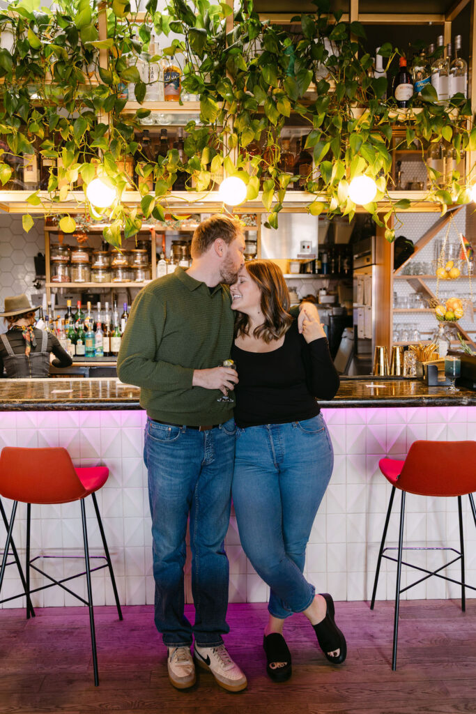 Couple leaning against the bar cuddling while the man gives the woman a gentle kiss on the forehead 