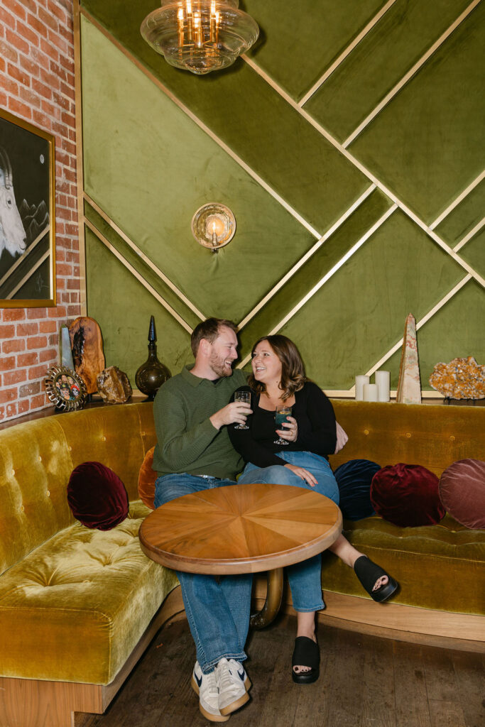 Couple cuddled on a velvet couch holding cocktails and sharing a candid moment during their engagement photos at a Denver cocktail bar 