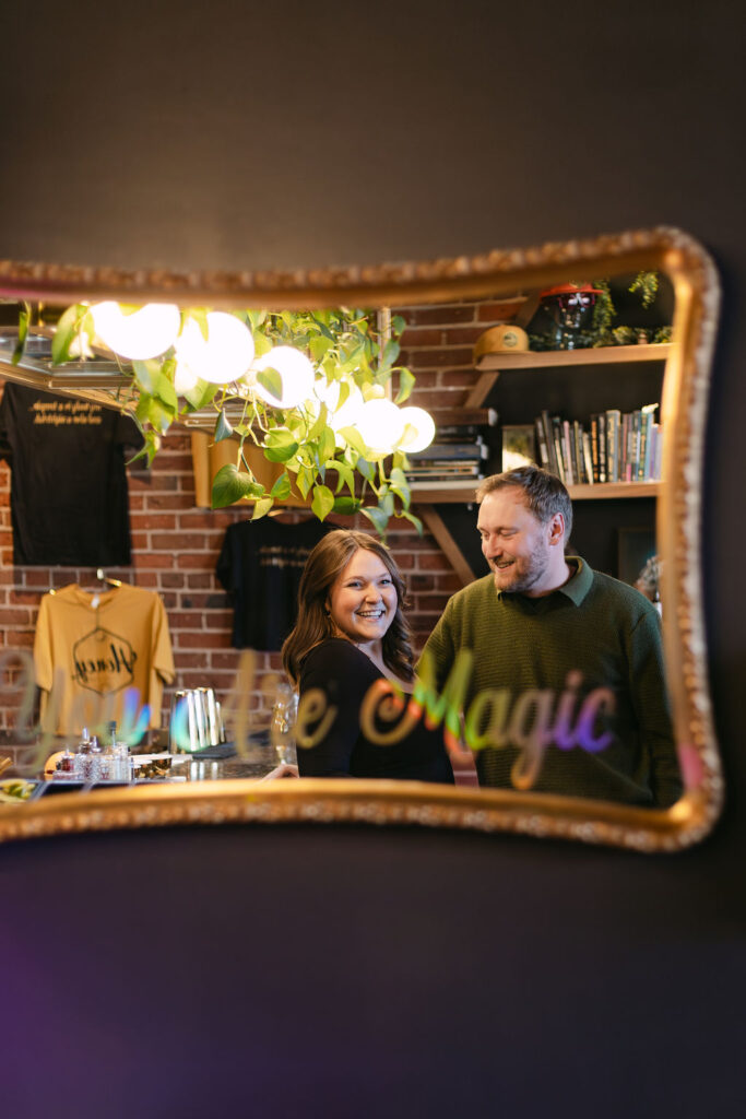 Reflection from a mirror of a girl smiling while her fiance stares lovingly at her leaning on the bar inside of Honey Elixir in Denver