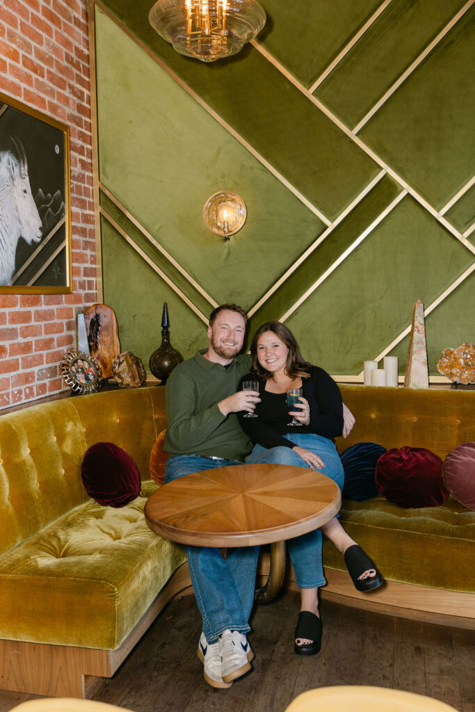 Couple lounging on a velvet couch during their engagement photos in Denver drinking cocktails and smiling