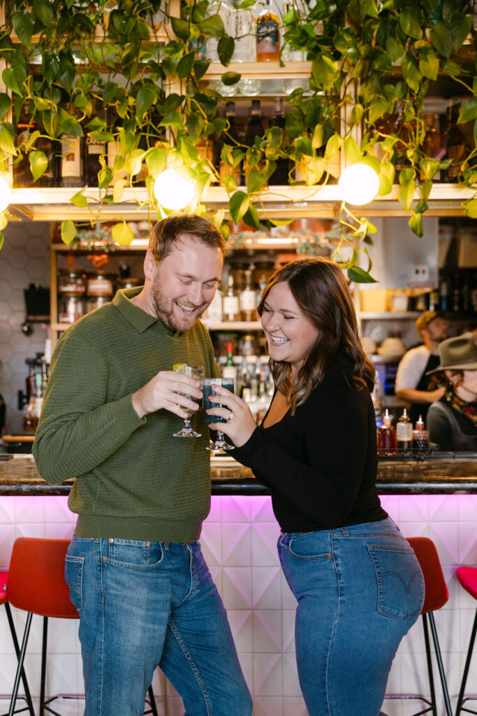 Candid engagement photo in a cocktail bar in denver with couple cheersing their colorful drinks while laughing