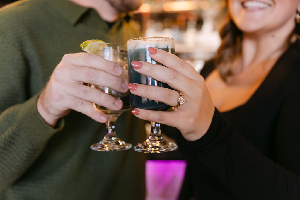 Close up image of a couple cheersing their colorful cocktails and the woman's engagement ring being showcased 