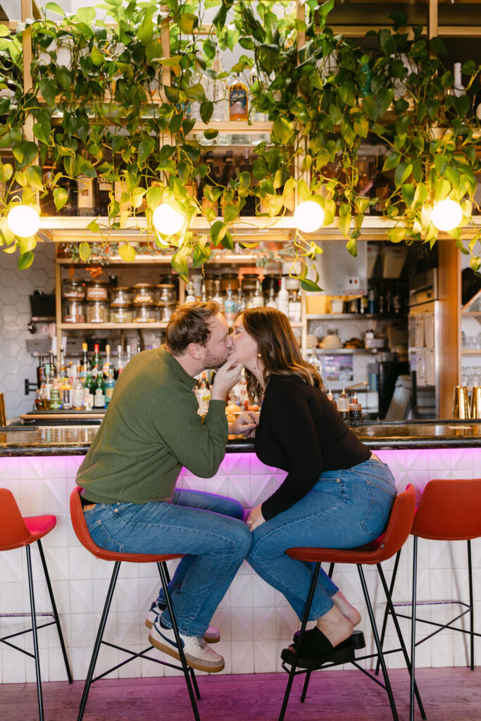 Couple sitting in red barstools at a vibrant bar, giving each other a kiss during their cocktail bar engagement photos in Denver