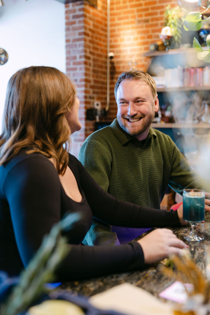 Natural engagement photos of a man laughing at his fiance sitting at the bar at Honey Elixir 