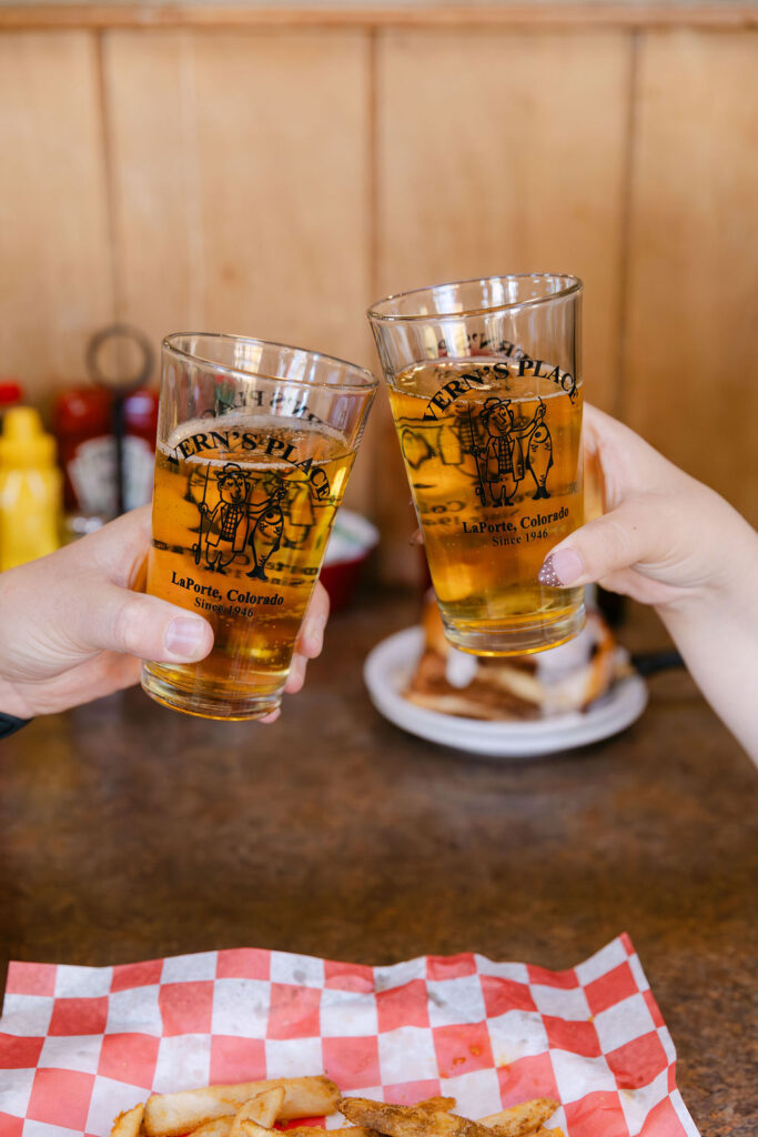 Hands holding up Vern’s Place drink glasses during a celebratory moment at their Colorado engagement session.