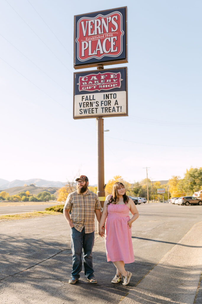 Full-length portrait of couple holding hands under the Vern’s Place sign with mountain views behind them during their Northern Colorado engagement photos