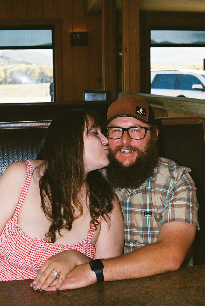 Couple sharing a sweet kiss on the cheek in a Vern’s Place booth during their Northern Colorado engagement photos