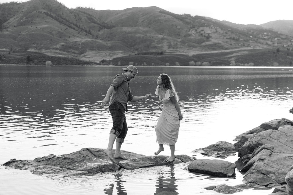 Black and white photo of Annelise and Ross holding hands while stepping across rocks in the water.